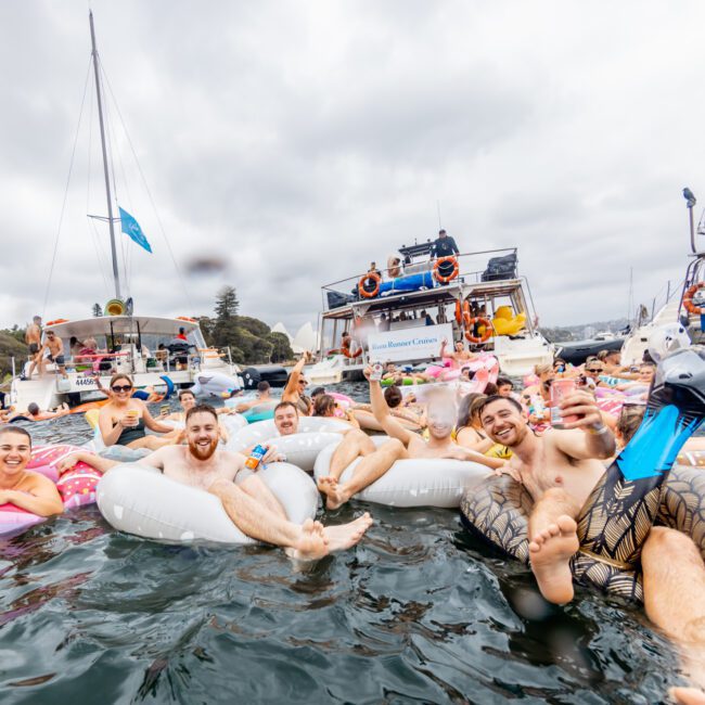 A lively group of people floating on inflatable rings in a large body of water, smiling and raising drinks. They are surrounded by several anchored boats, contributing to a festive atmosphere. Amidst the calm waters and overcast sky, this scene epitomizes The Yacht Social Club Sydney Boat Hire experience.