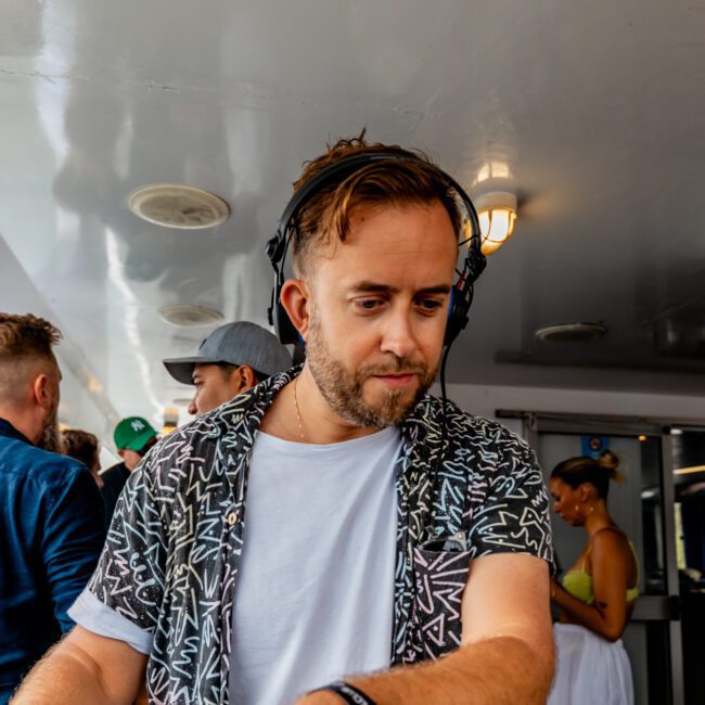 A DJ wearing headphones and a patterned shirt is mixing music at The Yacht Social Club on a luxurious yacht. People in the background are enjoying the event, with the stunning Sydney Harbour as their backdrop. The interior of the yacht, complete with atmospheric lighting and an open corridor, suggests a lively social gathering.