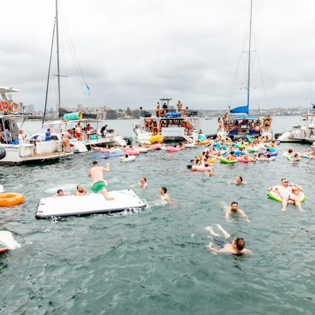 A lively scene unfolds as people enjoy The Yacht Social Club event on a lake, with several yachts anchored together. Many individuals swim and float on inflatables, while others gather and socialize on the boats under an overcast sky.