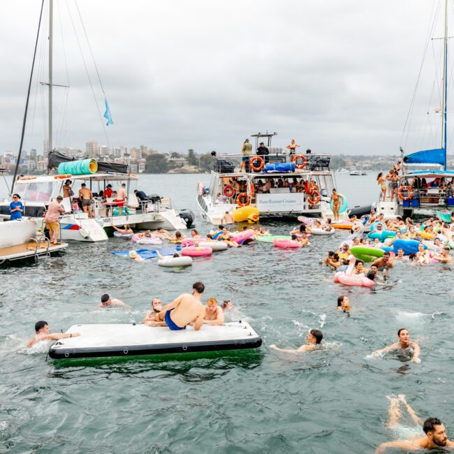 People enjoying a lively boat party on the water, surrounded by several boats and various inflatable floats. With swimmers and lounge-goers adding to the fun, an overcast sky looms above. The city skyline in the background hints at the vibrant atmosphere of Sydney Harbour Boat Hire The Yacht Social Club.