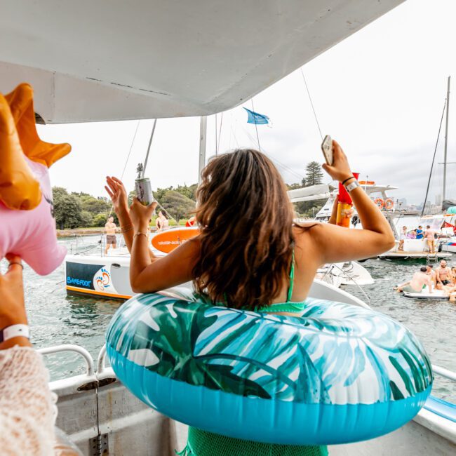 A person with a tropical-themed floaty ring around their waist stands on a boat, overlooking the water where several people are enjoying a float party. They hold their arms up, appearing to dance. Inflatable toys and distant trees are visible in the background, capturing the essence of The Yacht Social Club Event Boat Charters.