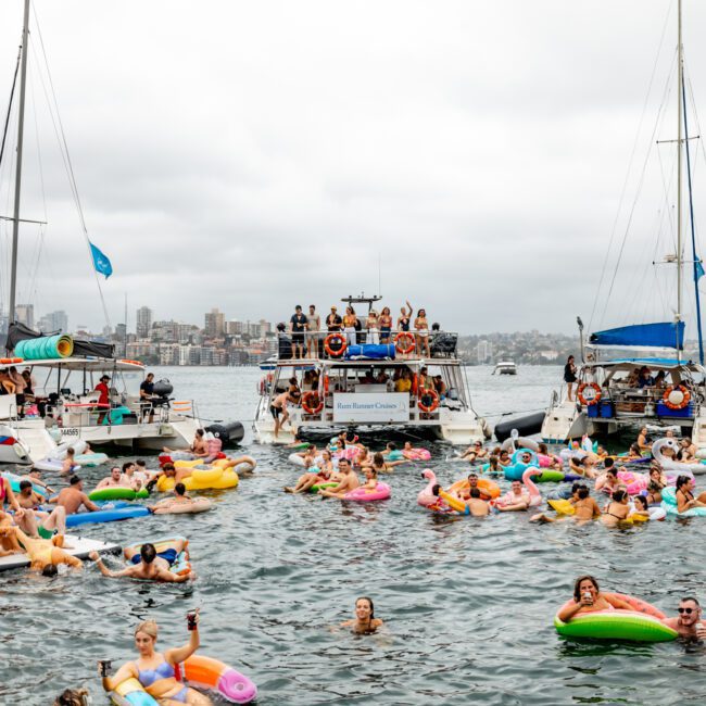 A vibrant scene on the water with numerous people floating on inflatable pool toys and swimming between several anchored boats. The background shows a city skyline under a cloudy sky. The atmosphere is lively and festive, reminiscent of events like The Yacht Social Club Sydney Boat Hire.