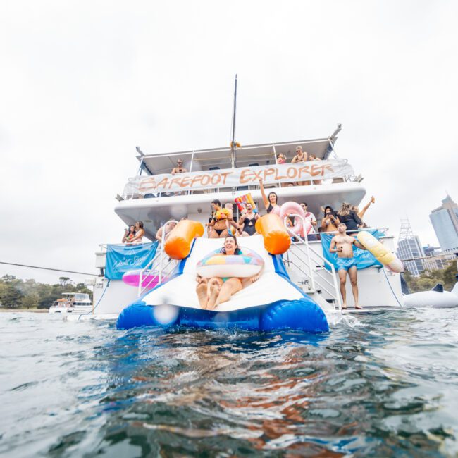 A large group of people on The Yacht Social Club's boat, "Barefoot Explorer," decorated with inflatable toys and banners. Docked on a calm body of water near a cityscape with tall buildings, people are enjoying themselves, some on the boat and others in the water.