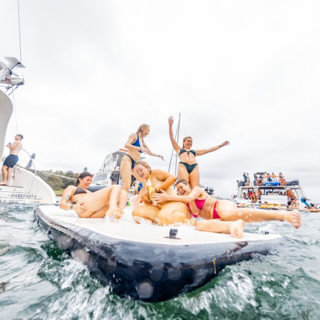 A group of people in swimsuits enjoying themselves on an inflatable raft on the water, surrounded by boats and other swimmers. The background features additional boats, cloudy skies, and individuals engaging in water activities, typical of a day with The Yacht Social Club Sydney Boat Hire.