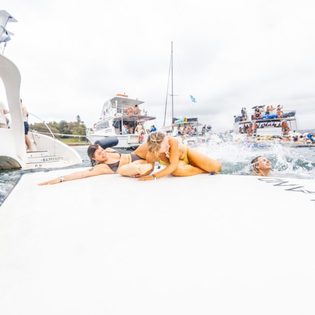 A lively scene of people enjoying a day on the water, featuring boats and a crowd of participants from The Yacht Social Club. The foreground shows individuals playfully lying on a large floating platform, adding to the festive and relaxed atmosphere of the gathering.