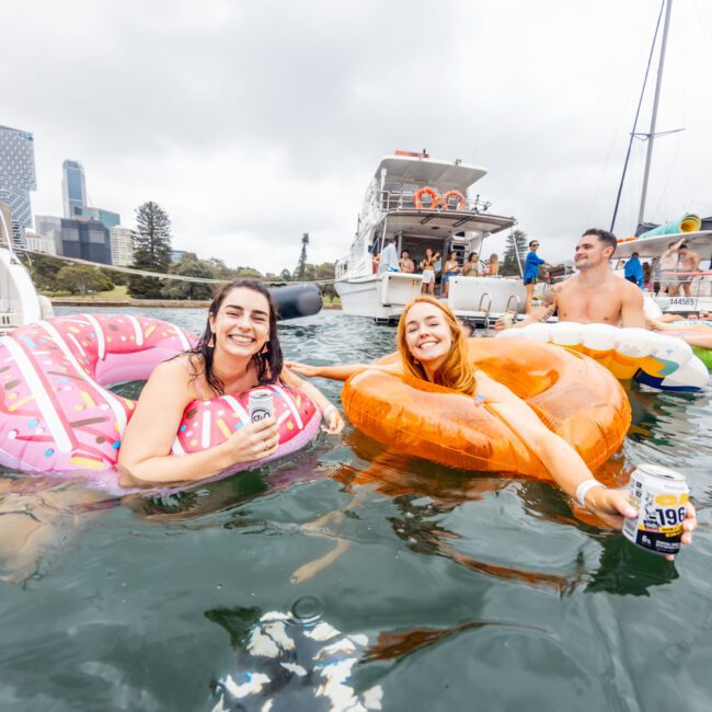 Two smiling women in colorful inflatable rings float in the water, holding cans of drinks. Behind them, people are on boats, including a large yacht and a small motorboat. Skyscrapers and trees are visible in the background with a cloudy sky above. Enjoy the scene with The Yacht Social Club Event Boat Charters.