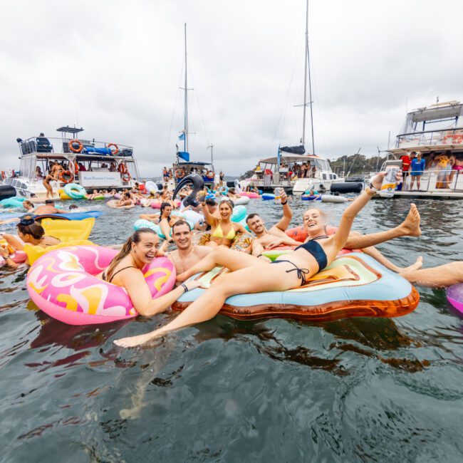 A lively group of people enjoying a party on various colorful inflatable floats in a body of water. Yachts are anchored nearby, and a cloudy sky sets the scene. People are laughing, smiling, and engaging in the festive atmosphere at The Yacht Social Club Event Boat Charters.
