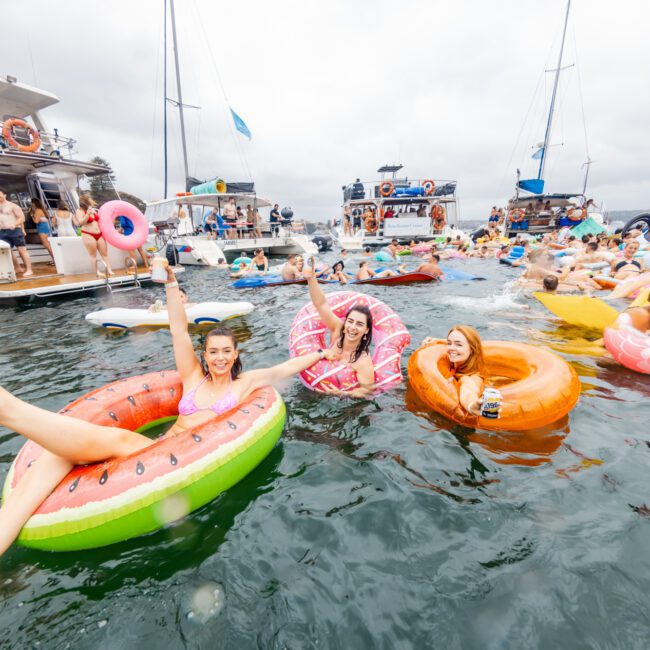 A lively scene of people enjoying a party on the water, sitting and floating on colorful inflatable pool toys shaped like a watermelon, donut, and flamingo. Various boats from The Yacht Social Club Sydney Boat Hire are anchored in the background with more partygoers. The atmosphere is festive and fun.