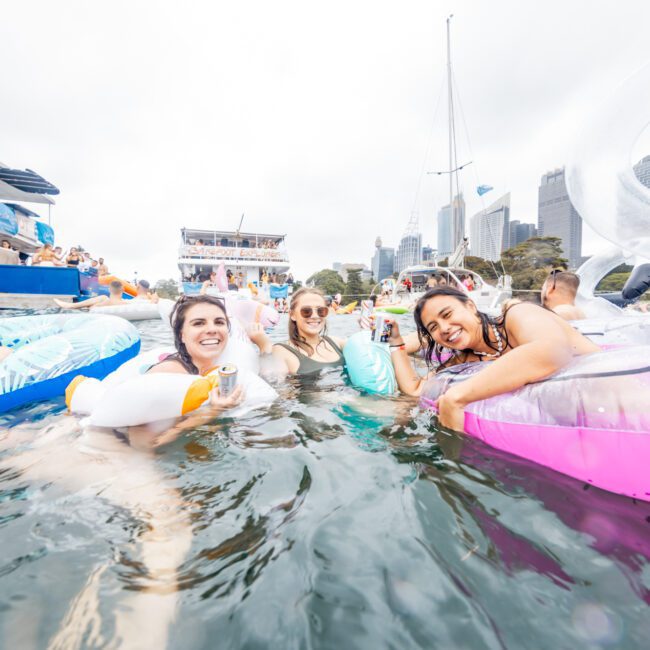 A group of smiling people enjoying themselves in the water, floating on various inflatable toys, including swans and tubes, near several boats. The background features a cityscape with tall buildings. A "Boat Parties Sydney The Yacht Social Club" logo appears in the bottom right corner.