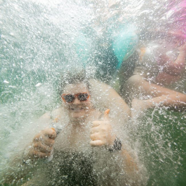 Underwater photo of people enjoying a splash in the water. The central figure wears red sunglasses and gives a double thumbs-up gesture, surrounded by bubbles. The image has a playful, energetic atmosphere. The logo "The Yacht Social Club Sydney Boat Hire" is visible in the bottom right corner.