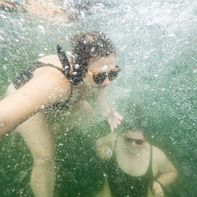 Two people in black swimsuits swim underwater in a green, bubbly environment. One person reaches out towards the other, both wearing sunglasses. The text "The Yacht Social Club - Boat Parties Sydney" appears in the lower right corner.