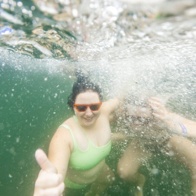 Two people submerged underwater are posing enthusiastically. One individual in a green swimsuit and orange sunglasses is giving a thumbs-up, embodying the fun of Boat Parties Sydney The Yacht Social Club. The water is bubbly and slightly murky, adding to the adventurous vibe. A watermark appears in the lower-right corner.
