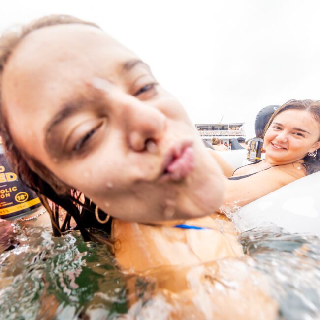 Two smiling people are relaxing in a pool or body of water. The person in the foreground is making a playful kissing face at the camera, while the person in the background smiles softly. Both are in flotation devices, with buildings visible in the distant background, reminiscent of a Luxury Yacht Rentals Sydney scene.