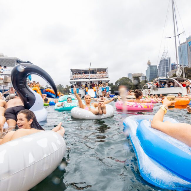 A lively scene of people enjoying a party on inflatable floaties, including swans and flamingos, in a marina with yachts and cityscape in the background. The festive atmosphere is enhanced by music and drinks. The image features "Yacht Social Club" branding, perfect for those considering Luxury Yacht Rentals Sydney.