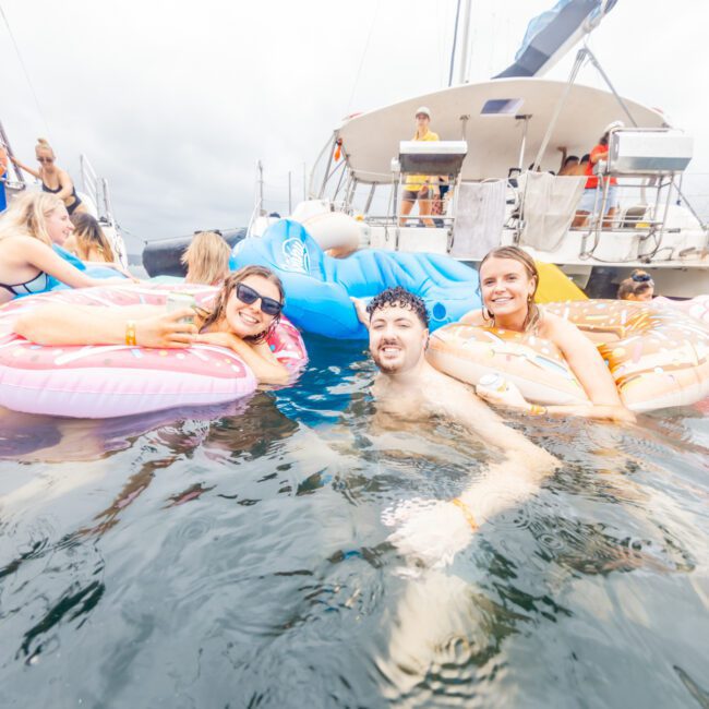 People are floating on inflatable pool toys in the water near a yacht. Some are lounging on pink and donut-shaped floats, while others stand or swim. The festive, casual atmosphere under a cloudy sky is part of "The Yacht Social Club Event Boat Charters." The Yacht Social Club logo is visible.