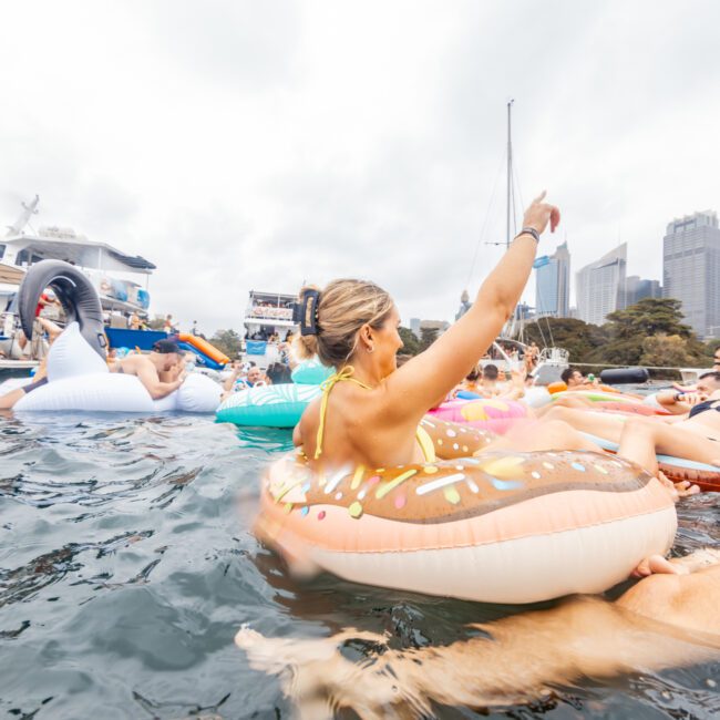 A group of people is enjoying a sunny day on the water, relaxing on various colorful inflatable floats, including a donut-shaped one. Luxury yachts and city skyscrapers are visible in the background. The atmosphere appears festive and lively, reminiscent of The Yacht Social Club Sydney Boat Hire experience.