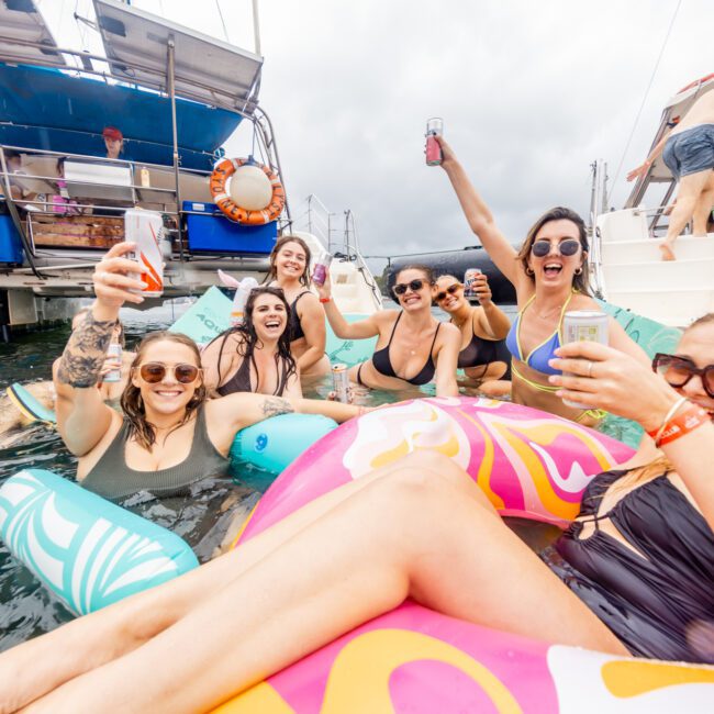 A group of friends enjoys a sunny day on the water, floating on colorful inflatables and holding drinks. Some are lounging on a boat while others are in the water, smiling and cheering. In the background, other boaters from The Yacht Social Club Sydney Boat Hire are seen on a nearby vessel.