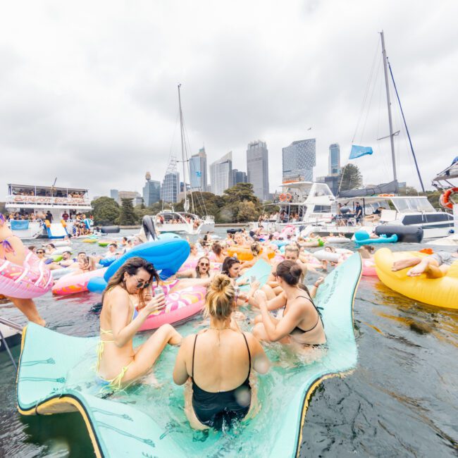 A group of people enjoys a fun day on the water, gathered on colorful inflatables and a large floating mat near docked boats. The backdrop features a city skyline with tall buildings under a cloudy sky. The lively scene feels like an event hosted by Sydney Harbour Boat Hire The Yacht Social Club.