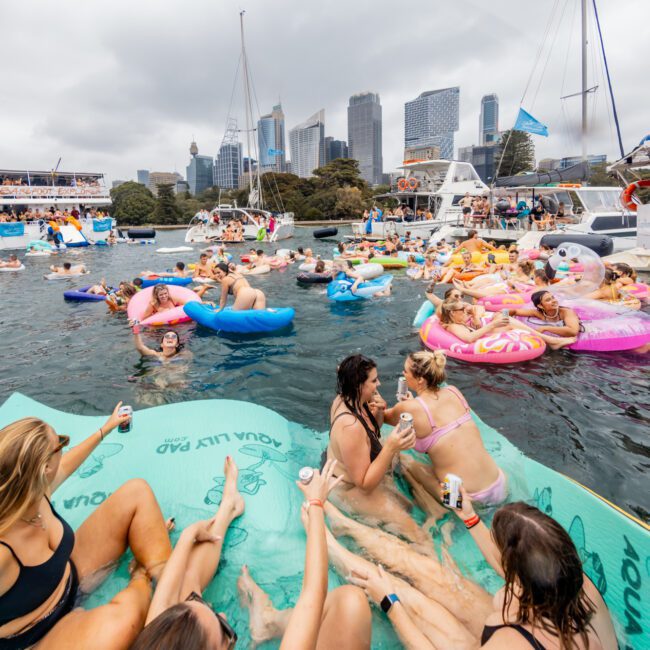 People are enjoying a lively boat party near a city skyline. Some are in the water on colorful floaties, while others relax on boats. The atmosphere is festive, with groups socializing and holding drinks. Buildings and trees are in the background, capturing the essence of The Yacht Social Club events.
