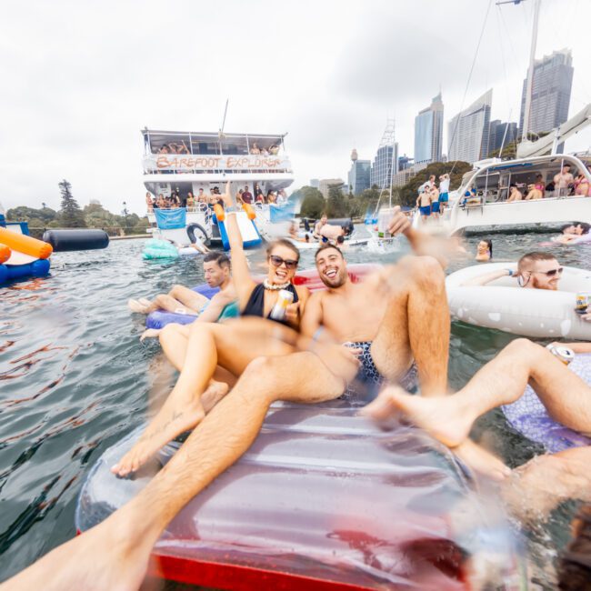A group enjoys a floating party on an inflatable raft in a body of water, surrounded by yachts and inflatable toys. Tall buildings are visible in the background. Some people are relaxing, while others engage in playful activities. It's the perfect scene for Sydney Harbour Boat Hire The Yacht Social Club.