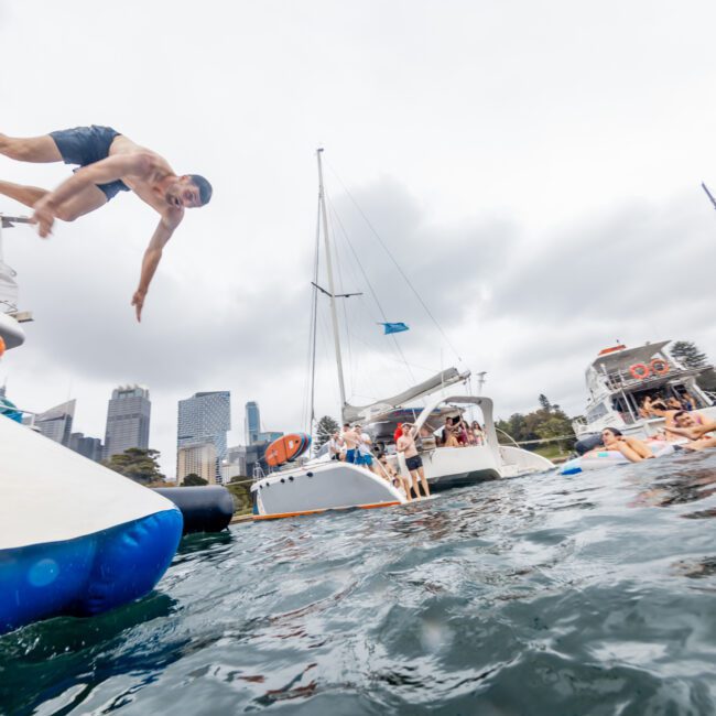 A man is captured mid-jump from a boat into the water during a lively gathering of boats hosted by The Yacht Social Club Sydney Boat Hire. Various people are seen relaxing on inflatable floats and aboard boats. The background features a city skyline under a cloudy sky, creating a fun, celebratory atmosphere.