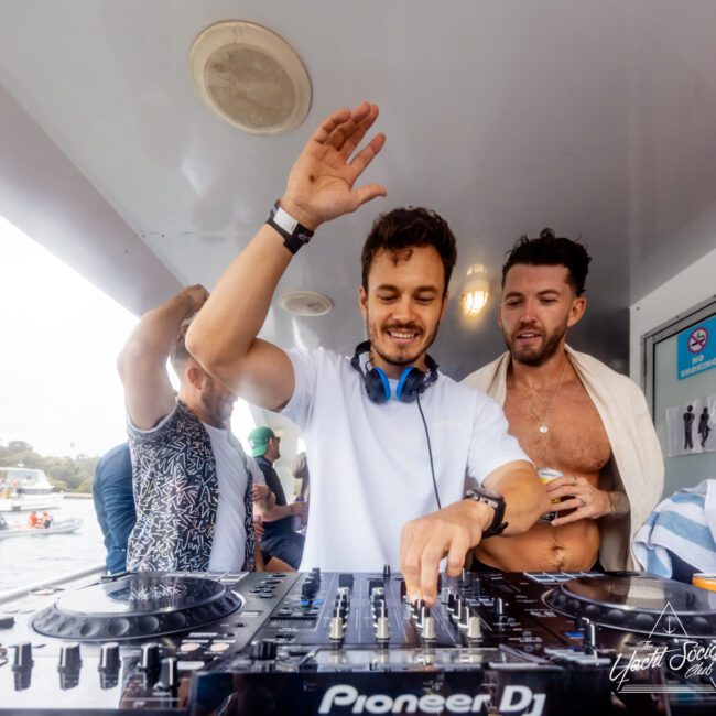 A DJ in a white t-shirt and headphones around his neck is mixing music on a Pioneer DJ console. He is inside a boat from The Yacht Social Club Sydney Boat Hire, surrounded by four people. One person stands close, partially draped in a towel. The atmosphere is lively, perfect for parties.