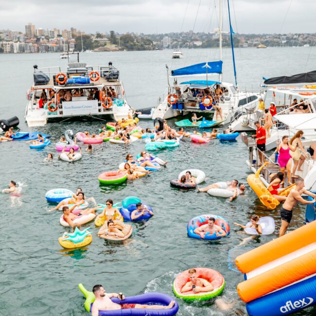 A lively gathering of people enjoying a sunny day on the water, floating on various colorful inflatable rafts and boats near anchored yachts. The background features a city skyline under a slightly overcast sky. A "The Yacht Social Club" sign is seen in the bottom right, exemplifying Sydney Harbour Boat Hire.