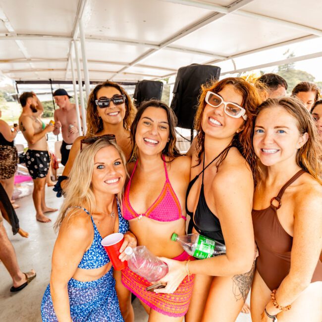 A group of people enjoying a boat party. Five women in swimwear are posing for the camera in the foreground, smiling and holding drinks. The background shows several people socializing and dancing on the deck under a canopy. The atmosphere is lively and festive at Boat Parties Sydney The Yacht Social Club.
