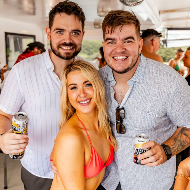 Three people smile while posing for a photo on a boat. The woman in front wears a red bikini, while the two men behind her wear short-sleeved shirts and hold cans of beverages. A disco ball hangs from the ceiling, and other passengers are in the background enjoying The Yacht Social Club Event Boat Charters.