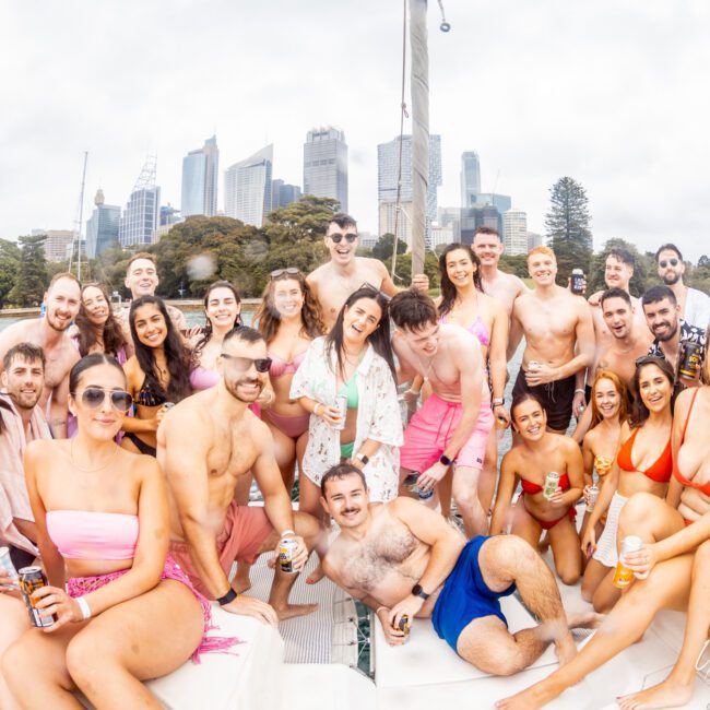 A large group of people in swimwear is gathered on a boat, smiling and posing for the camera. They seem to be enjoying a day out on the water organized by The Yacht Social Club Sydney Boat Hire. The city skyline with tall buildings and some greenery are visible in the background under a cloudy sky.