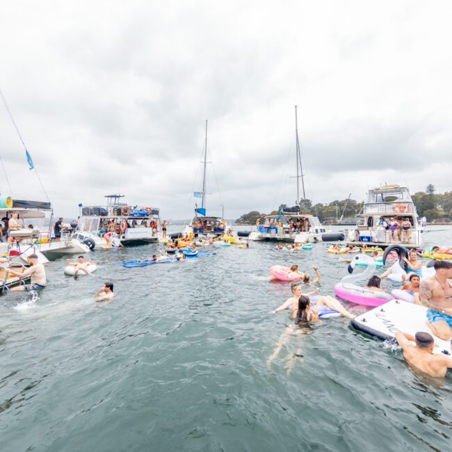 A lively scene on the water with numerous people swimming and floating on various inflatables. Several boats and yachts from The Yacht Social Club Sydney Boat Hire are anchored nearby, each filled with more people enjoying the day. The sky is overcast, and the atmosphere appears festive and fun.