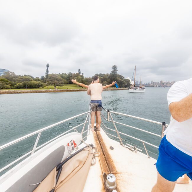 A man stands on the bow of a boat with his arms outstretched facing a body of water, while another man drinks from a cup on the right. Trees and buildings frame the background. The logo "The Yacht Social Club" appears in the bottom right corner, highlighting Luxury Yacht Rentals Sydney.