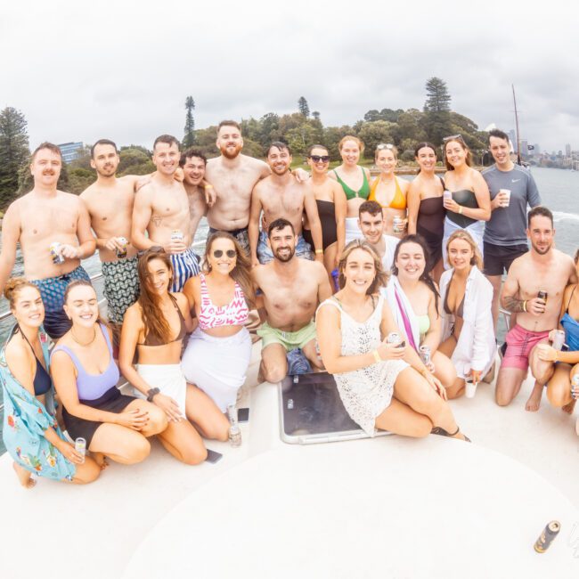 A group of people stands and sits on a boat, with water and a partial city skyline in the background. The group, dressed in casual and swimwear, smiles at the camera. The surroundings include trees and a cloudy sky—an ideal scene for The Yacht Social Club Event Boat Charters.