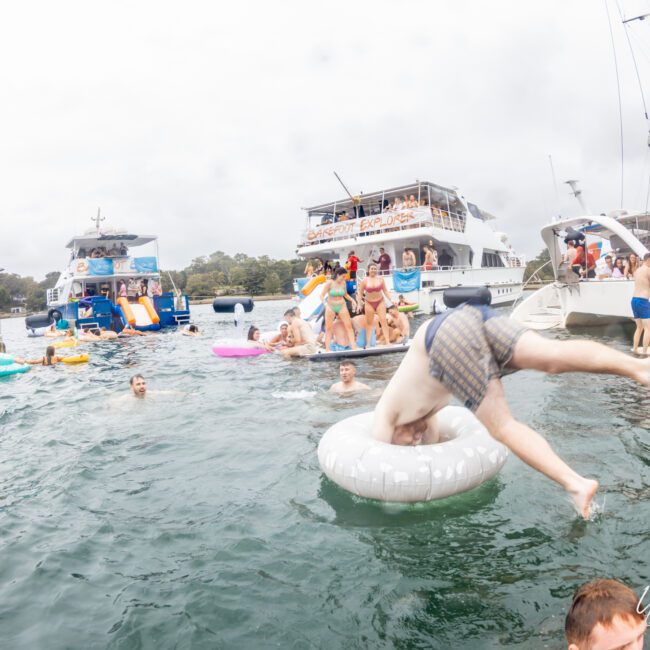 A lively scene of people enjoying a day on the water. Several boats from The Yacht Social Club are docked nearby, with individuals on inflatables and swimming in the water. In the foreground, a person is mid-flip into a white inflatable ring. The background shows trees and cloudy skies.