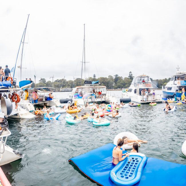 People are socializing and relaxing on various floating devices in a lively, crowded marina. Several boats from Sydney Harbour Boat Hire The Yacht Social Club are docked nearby, and many participants are seen enjoying the sunny day on the water. A light-colored inflatable platform is positioned in the center.