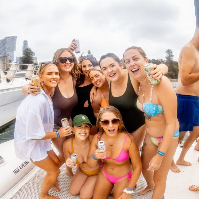 A group of friends, mostly women, wearing swimsuits are posing and smiling on a boat docked by a city skyline. They are holding drinks and seem to be enjoying a sunny day at The Yacht Social Club Sydney Boat Hire. Other people can be seen in the background, also having a good time.