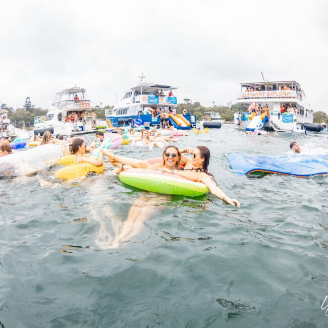 A group of people enjoy a sunny day on the water, floating on inflatable tubes, and playing on a water mat. Several yachts from The Yacht Social Club are anchored in the background, with more individuals socializing on board. The atmosphere is festive and lively.