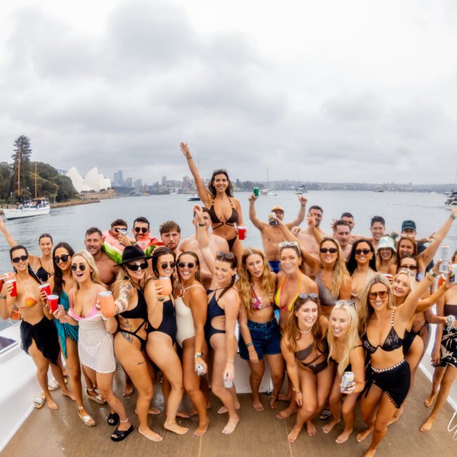 A group of people in swimsuits are gathered on a boat, posing and smiling for a photo. They hold drinks and appear to be celebrating. The Yacht Social Club Sydney Boat Hire provides the perfect backdrop with a cloudy sky, water with a sailboat, distant buildings, and trees in the background.