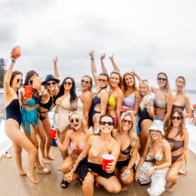 A group of 15 women pose and smile on the deck of a yacht. They are wearing swimsuits, sunglasses, and holding drinks. The background shows an overcast sky and water. A logo in the bottom right corner reads "Luxury Yacht Rentals Sydney." Everyone appears cheerful and relaxed at The Yacht Social Club.