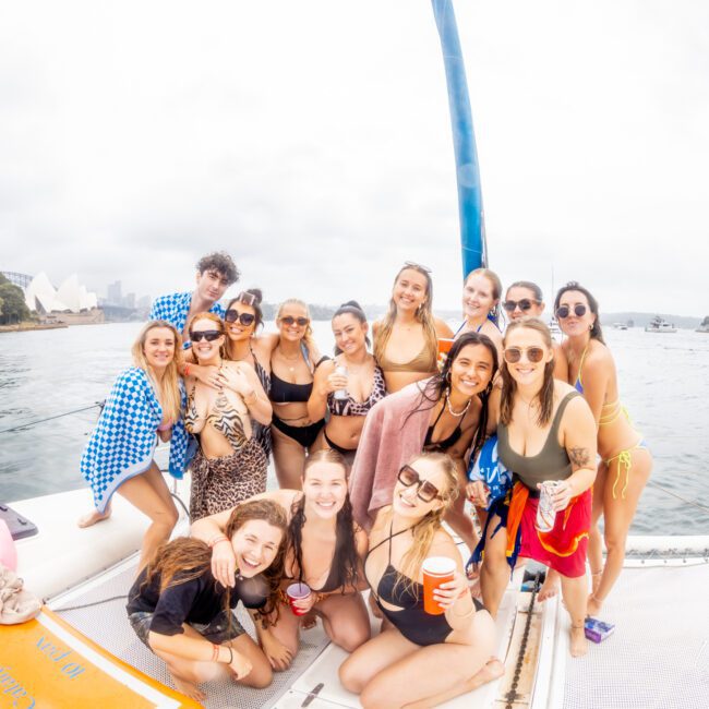 A group of fifteen young adults, mostly women, are gathered on a boat, posing for a photo. They are wearing swimwear and have drinks in hand. The background shows a cloudy sky, calm water, and some trees and boats in the distance. Everyone is smiling and enjoying the moment at Boat Parties Sydney The Yacht Social Club.
