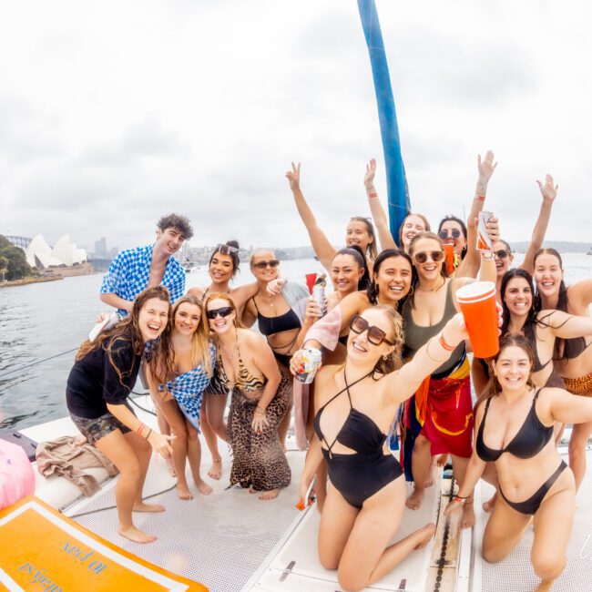 A group of people in swimsuits are posing and celebrating on a boat from The Yacht Social Club Sydney Boat Hire with drinks in their hands. They are smiling and enjoying a festive boat party. The background shows water, a coastline with trees, and a distant architectural structure.
