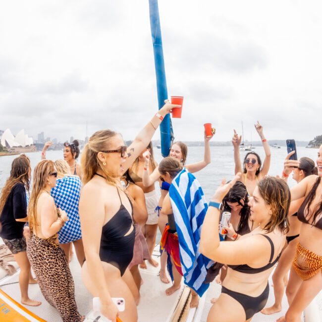 A group of people in swimwear are having a lively party on a boat from Boat Parties Sydney The Yacht Social Club. They are holding red cups, dancing, and laughing. The background features a body of water with sailboats and a city skyline partially visible under a cloudy sky.
