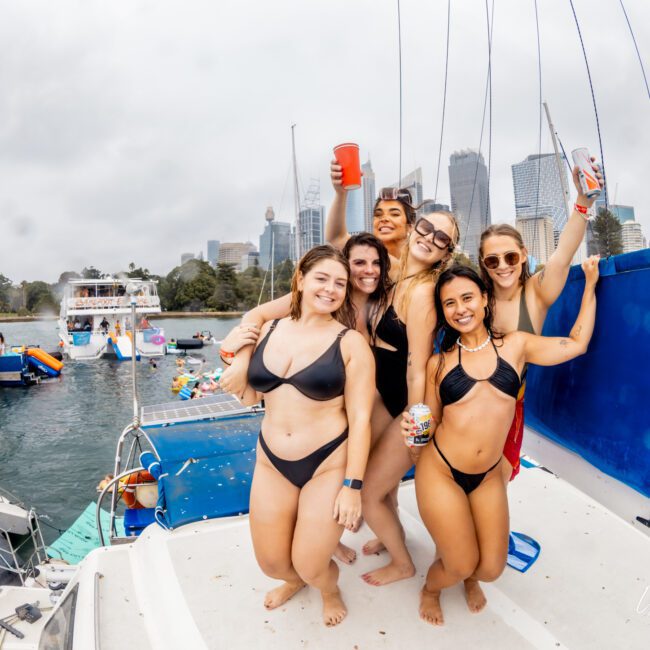 Six women in swimsuits stand close together on a boat, smiling and holding drinks while posing for a photo at The Yacht Social Club Event Boat Charters. Other boats and people are visible in the background on the water with a city skyline under a cloudy sky.
