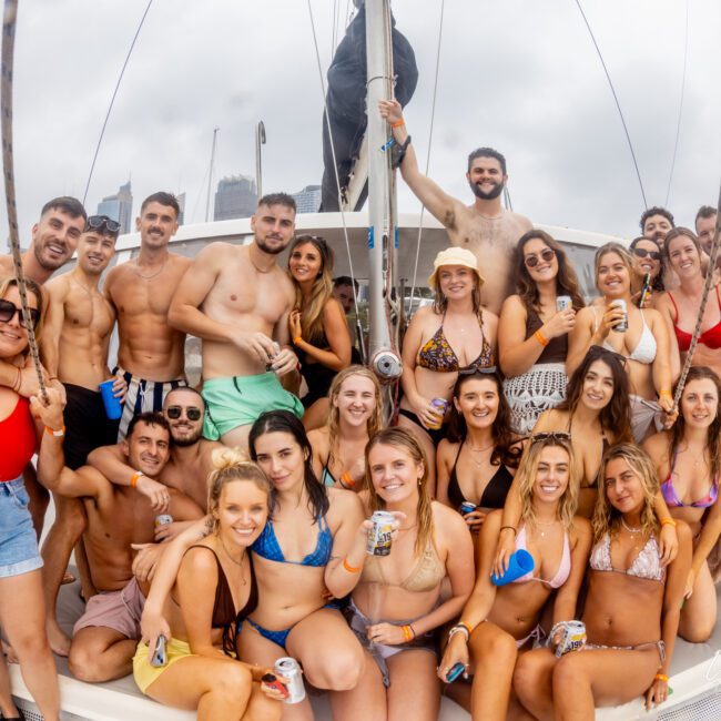 A group of people in swimsuits stand closely together on a boat, smiling and holding drinks. The background shows other boats and a cloudy sky. The scene is lively with a sense of fun and camaraderie among the group, typical of an outing with The Yacht Social Club Sydney Boat Hire.