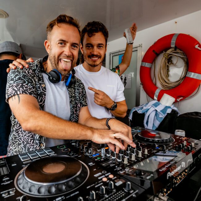 Two men are standing behind DJ equipment on a boat. The man on the left, sticking out his tongue while using the controls, wears headphones and a patterned shirt. The man on the right smiles and points at him. A lifebuoy is visible in the background, showcasing fun with Sydney Harbour Boat Hire The Yacht Social Club.