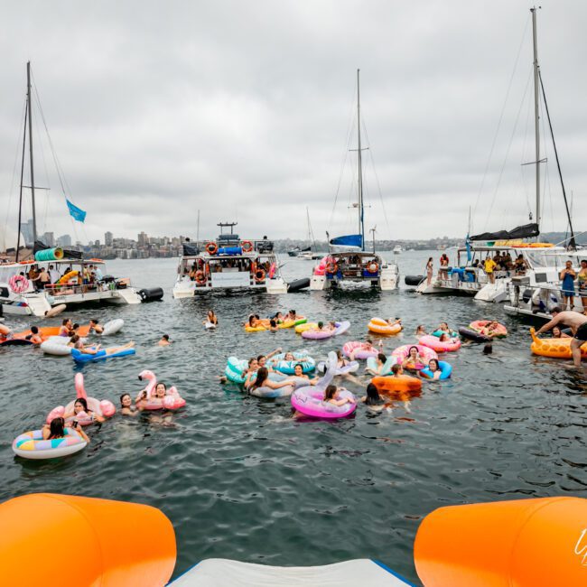 People float on various inflatable rafts, lounging and socializing in the water near several docked boats on a cloudy day. The scene is festive, with a mix of colorful floaties and a few people swimming. A logo in the bottom right corner reads "Sydney Harbour Boat Hire The Yacht Social Club".