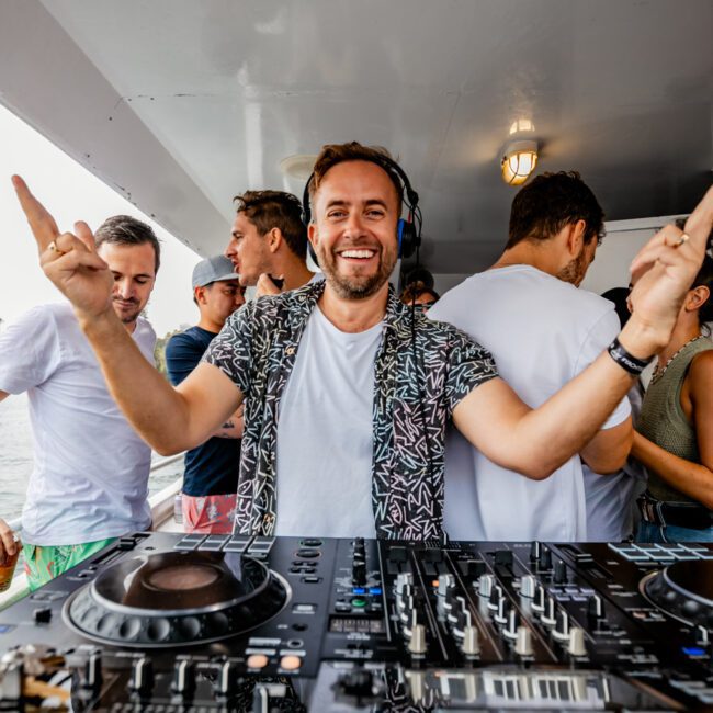 A DJ wearing headphones and a patterned shirt smiles energetically while standing behind a DJ console on a boat. People in casual attire are gathered around him, enjoying the event. The scene is lively and festive, with "The Yacht Social Club" visible in the corner, exemplifying Luxury Yacht Rentals Sydney.