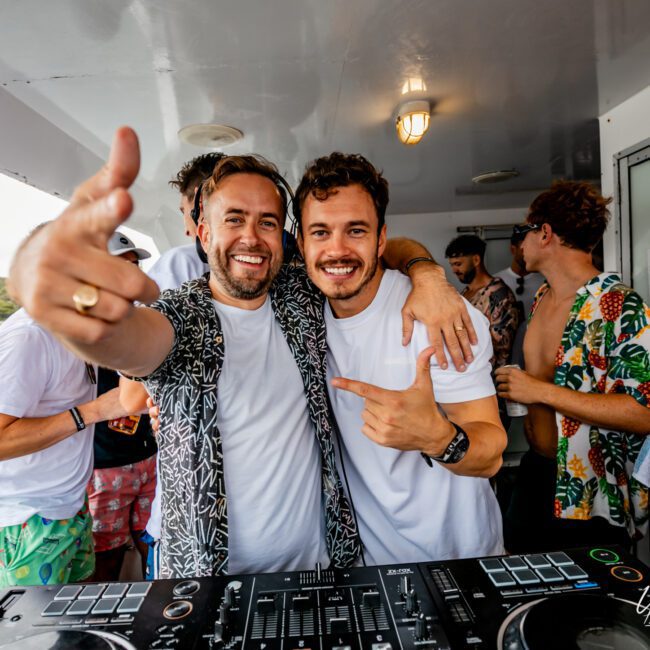 Two men stand side by side, smiling and pointing at the camera on a boat with friends. The man on the left wears a floral shirt, while the one on the right sports a white shirt. In the foreground, there is DJ equipment. Enjoy your time with The Yacht Social Club Sydney Boat Hire for unforgettable boat parties!