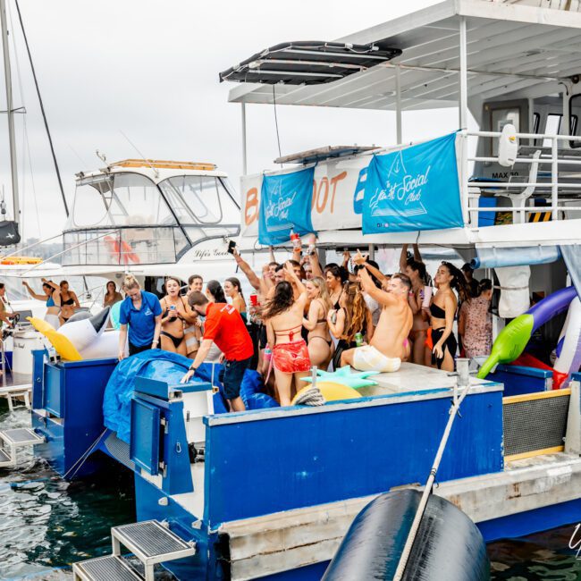 A large group of people in swimwear are partying on two yachts docked side by side in the water. Some are dancing, others are cheering and taking photos. The yacht in the foreground has a banner reading "The Yacht Social Club Sydney Boat Hire." Inflatable toys can be seen around the boats.