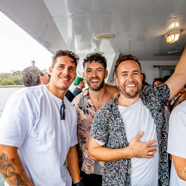 Three men standing closely together on a boat, all smiling towards the camera. The man on the left is wearing a white t-shirt, the man in the middle is in a patterned shirt, and the man on the right is in a dark patterned shirt with a white t-shirt underneath. Water and trees are in the background, enjoying Sydney Harbour Boat Hire from The Yacht Social Club.